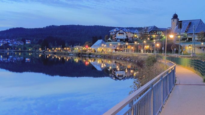 Ruhevoller See mit beleuchteter Uferpromenade und sanften Hügeln im Hintergrund.