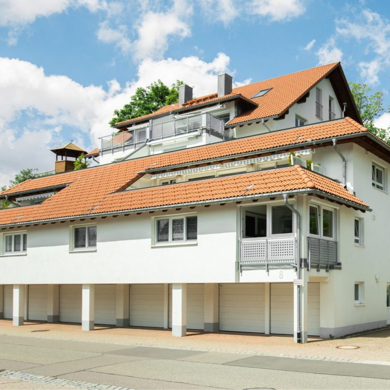 Weißes, mehrstöckiges Haus mit roten Dachziegeln und ebenerdigen Garagen.
