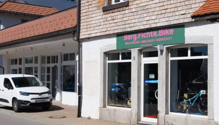 Fahrradshop mit Service und Fahrradverleih rechts von der Ferienwohnung, links neben dem Shop ein weißes Auto vor einer Schaufensterfront.
