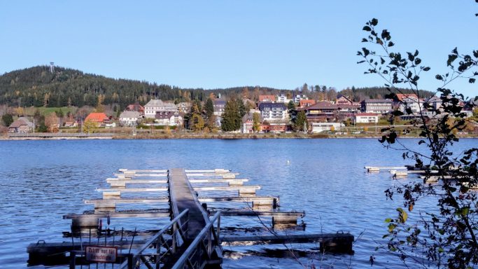 Blick auf ein Gewässer mit Steg, umgeben von Hügeln und einem kleinen Dorf.