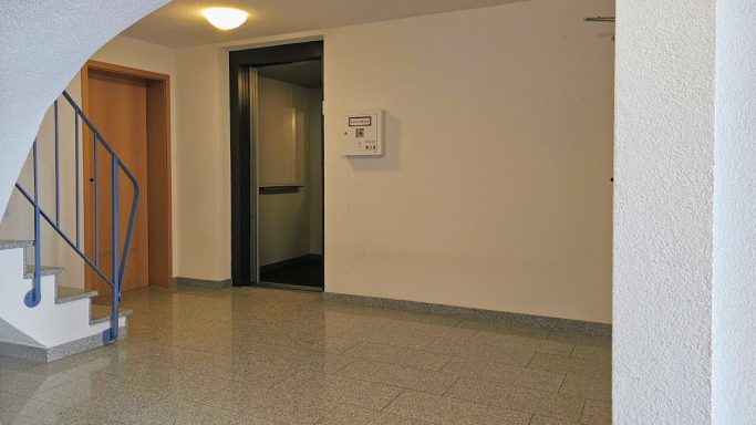Flur mit Treppe, Aufzugstür und weißen Wänden. Boden aus grauem Fliesenmaterial.