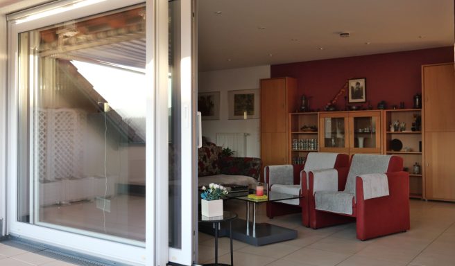 Blick von der Terrasse auf das Wohnzimmer. Vorne 2 Sessel mit Couchtisch, rechts Schränke